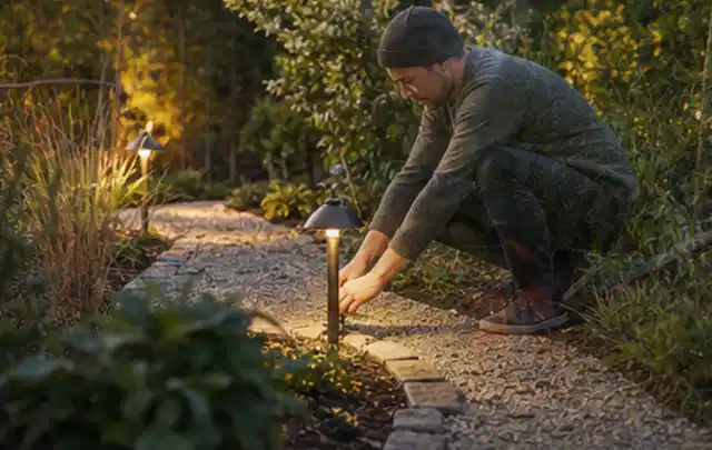 Person richtet eine niedrige warm leuchtende Gartenlampe am Kiesweg aus; Thema Gartenwerkstatt, Schwerpunkt Gartenhäuser; Kontrolle.