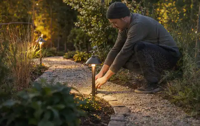 Person richtet eine niedrige warm leuchtende Gartenlampe am Kiesweg aus; Thema Gartenwerkstatt, Schwerpunkt Nachhaltigkeit; Umsetzung.