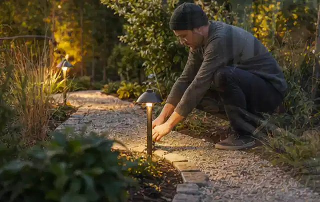 Person richtet eine niedrige warm leuchtende Gartenlampe am Kiesweg aus; Thema Gartenwerkstatt, Schwerpunkt Pflege; Ausgangslage.