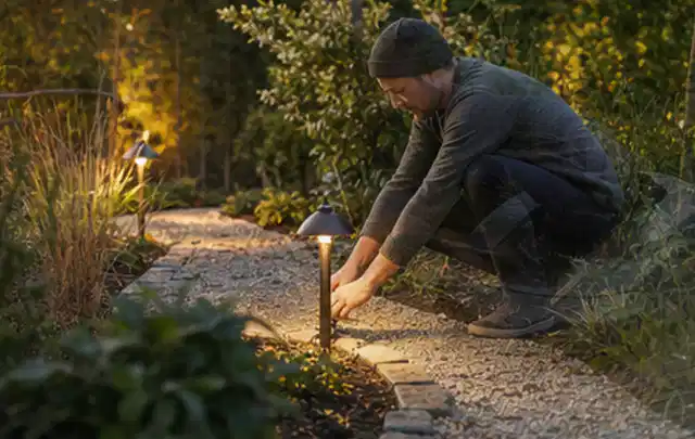 Person richtet eine niedrige warm leuchtende Gartenlampe am Kiesweg aus; Thema Gartenwerkstatt, Schwerpunkt Planung; Umsetzung.