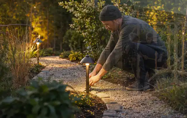 Person richtet eine niedrige warm leuchtende Gartenlampe am Kiesweg aus; Thema Gasgrills, Schwerpunkt Bestenliste; Kontrolle.