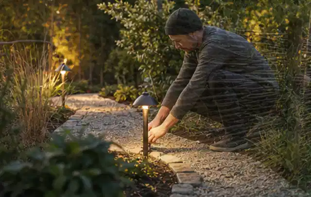 Person richtet eine niedrige warm leuchtende Gartenlampe am Kiesweg aus; Thema Gasgrills, Schwerpunkt Dimensionierung; Ergebnis.