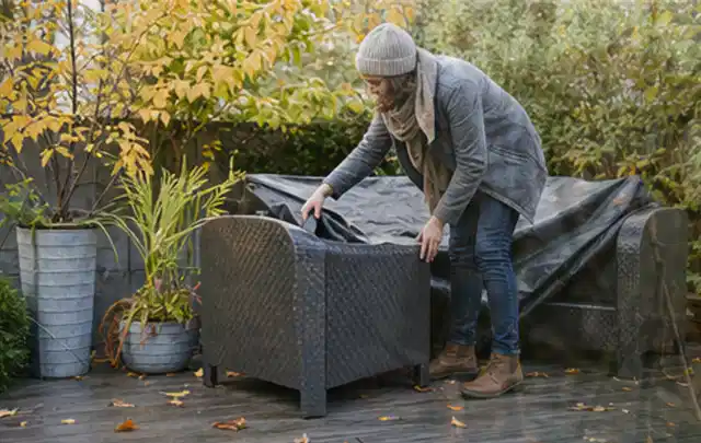 Person deckt Gartenmöbel vor der kalten Jahreszeit sorgfältig ab; Thema Gasgrills, Schwerpunkt Dimensionierung; Praxisalltag.