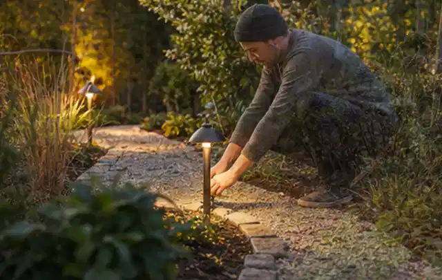 Person richtet eine niedrige warm leuchtende Gartenlampe am Kiesweg aus; Thema Gasgrills, Schwerpunkt Ersatzteile; Praxisalltag.
