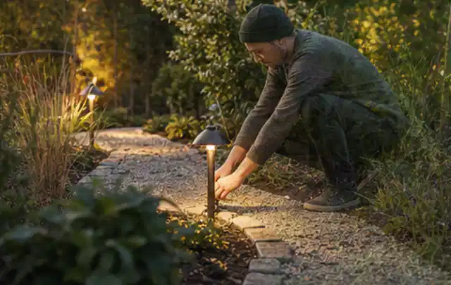 Person richtet eine niedrige warm leuchtende Gartenlampe am Kiesweg aus; Thema Gasgrills, Schwerpunkt Fehlerhilfe; Praxisalltag.