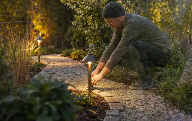 Person richtet eine niedrige warm leuchtende Gartenlampe am Kiesweg aus; Thema Gasgrills, Schwerpunkt Gärten mit kleiner Fläche; Praxisalltag.