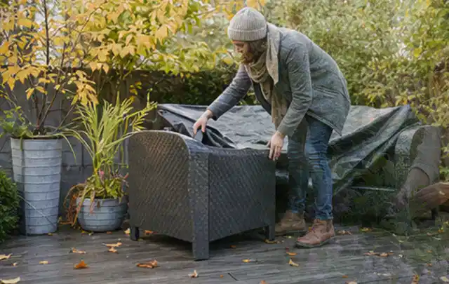 Person deckt Gartenmöbel vor der kalten Jahreszeit sorgfältig ab; Thema Gasgrills, Schwerpunkt Reinigung; Praxisalltag.