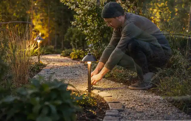 Person richtet eine niedrige warm leuchtende Gartenlampe am Kiesweg aus; Thema Gasgrills, Schwerpunkt Reparatur; Umsetzung.