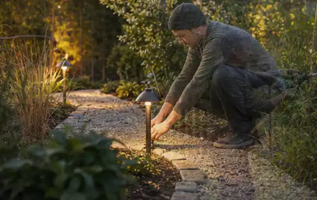 Person richtet eine niedrige warm leuchtende Gartenlampe am Kiesweg aus; Thema Geräteschuppen, Schwerpunkt Aufbau; Ausgangslage.