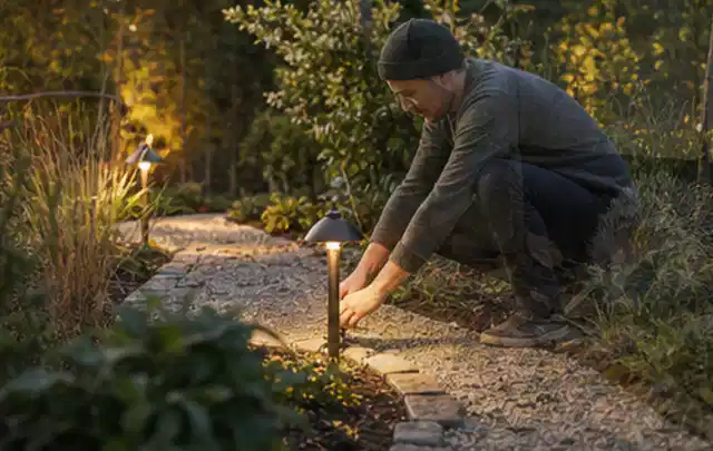 Person richtet eine niedrige warm leuchtende Gartenlampe am Kiesweg aus; Thema Geräteschuppen, Schwerpunkt Baurecht; Praxisalltag.