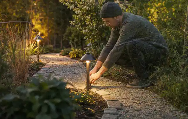 Person richtet eine niedrige warm leuchtende Gartenlampe am Kiesweg aus; Thema Geräteschuppen, Schwerpunkt Gärten mit kleiner Fläche; Praxisalltag.