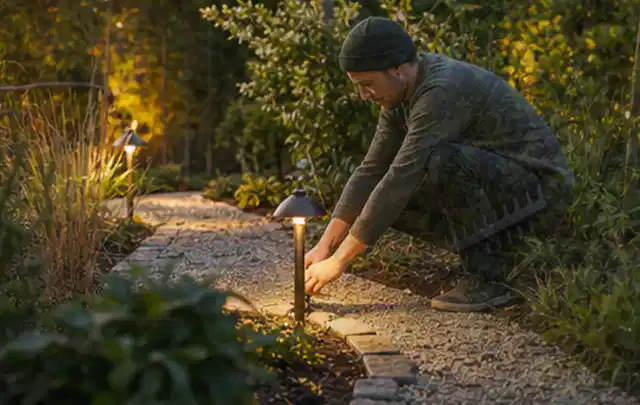 Person richtet eine niedrige warm leuchtende Gartenlampe am Kiesweg aus; Thema Geräteschuppen, Schwerpunkt Winterschutz; Umsetzung.