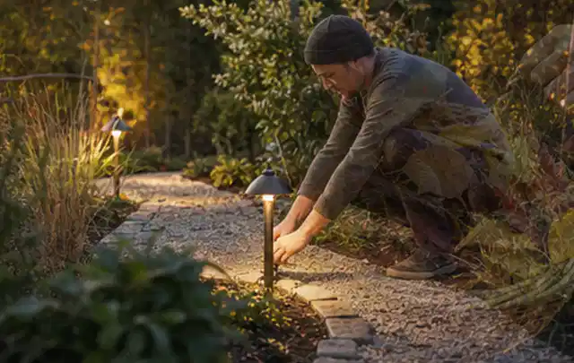Person richtet eine niedrige warm leuchtende Gartenlampe am Kiesweg aus; Thema Hanggarten, Schwerpunkt Anleitung; Umsetzung.