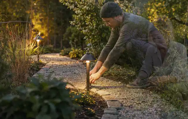 Person richtet eine niedrige warm leuchtende Gartenlampe am Kiesweg aus; Thema Hanggarten, Schwerpunkt Praxisbeispiele; Ausgangslage.