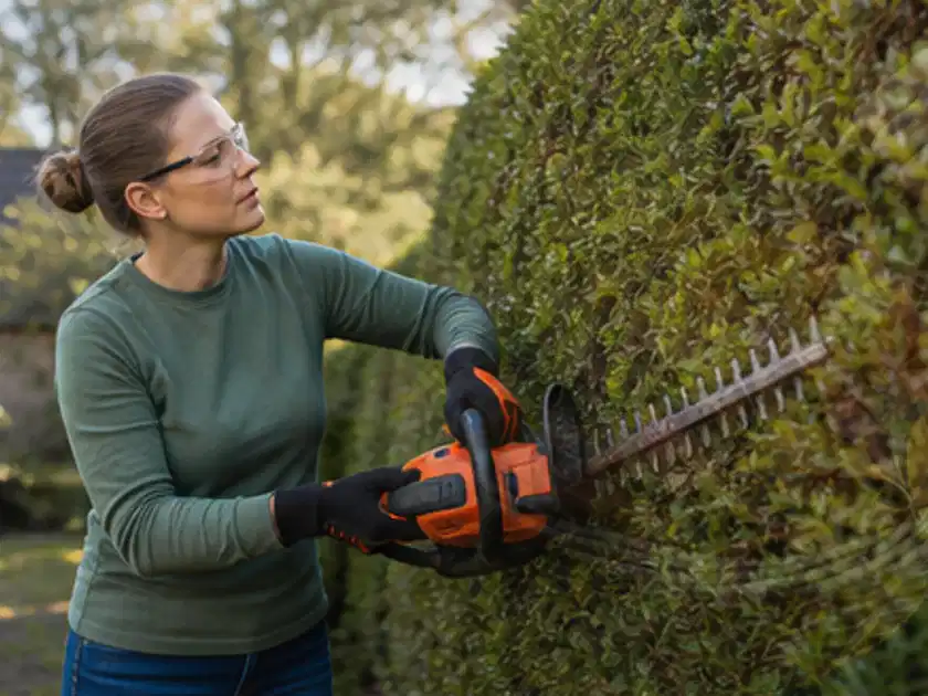 Person schneidet eine dichte Gartenhecke mit Schutzbrille und sicherem Stand; Thema Heckenscheren, Schwerpunkt Ersatzteile; Titelbild.