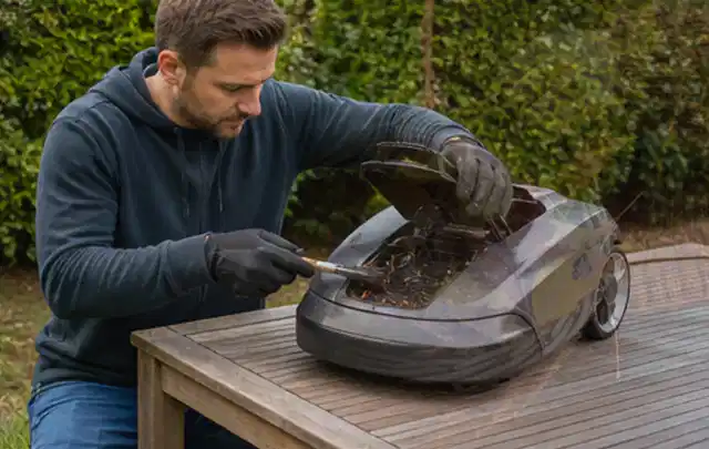 Person wartet einen geöffneten Mähroboter auf einem stabilen Gartentisch; Thema Heckenscheren, Schwerpunkt Kosten; Ausgangslage.