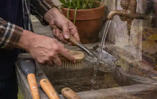 Hände reinigen gebrauchte Gartengeräte mit Bürste und Wasser am Außenbecken; Thema Heckenscheren, Schwerpunkt Systemvergleich; Umsetzung.