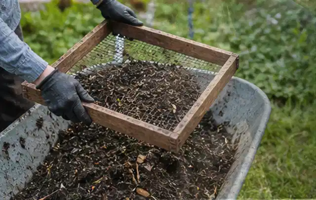Behandschuhte Hände sieben reifen Kompost über einer Gartenschubkarre; Thema Hochbeet Pflege, Schwerpunkt Checkliste; Umsetzung.