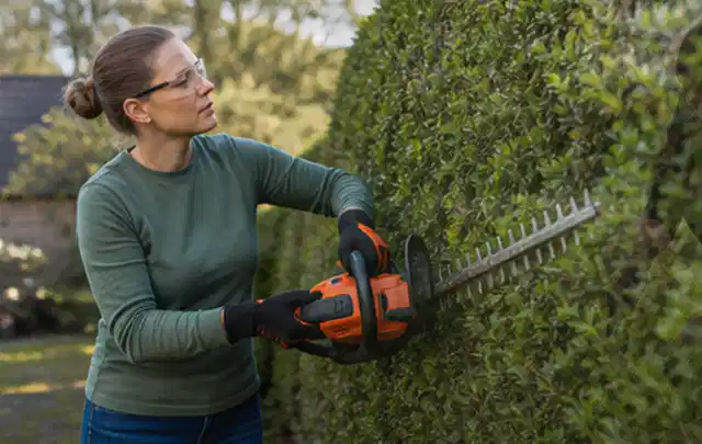 Person schneidet eine dichte Gartenhecke mit Schutzbrille und sicherem Stand; Thema Hochdruckreiniger, Schwerpunkt Ersatzteile; Praxisalltag.