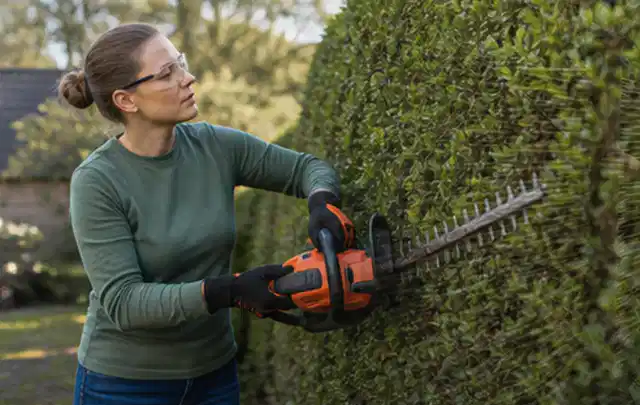 Person schneidet eine dichte Gartenhecke mit Schutzbrille und sicherem Stand; Thema Hochdruckreiniger, Schwerpunkt Gartengeräte; Kontrolle.