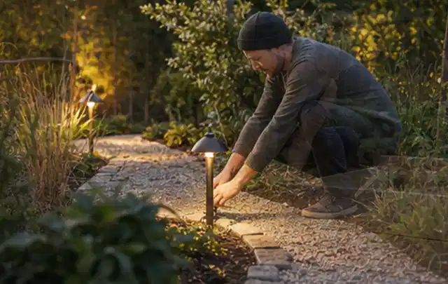 Person richtet eine niedrige warm leuchtende Gartenlampe am Kiesweg aus; Thema Hollywoodschaukeln, Schwerpunkt Ersatzteile; Umsetzung.