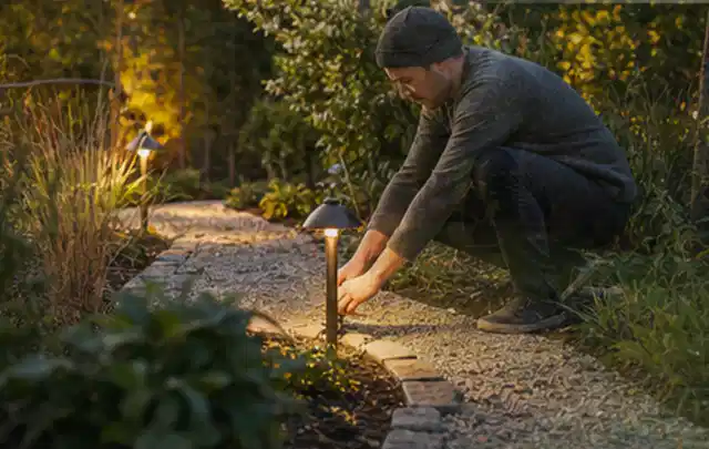 Person richtet eine niedrige warm leuchtende Gartenlampe am Kiesweg aus; Thema Hollywoodschaukeln, Schwerpunkt Gärten mit großer Fläche; Ergebnis.
