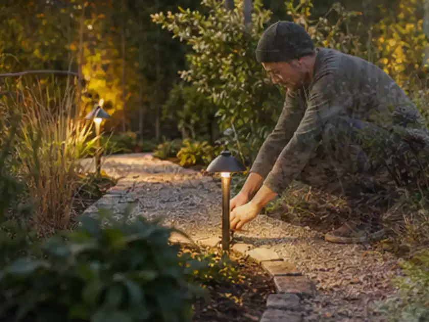 Person richtet eine niedrige warm leuchtende Gartenlampe am Kiesweg aus; Thema Hollywoodschaukeln, Schwerpunkt Gärten mit kleiner Fläche; Titelbild.