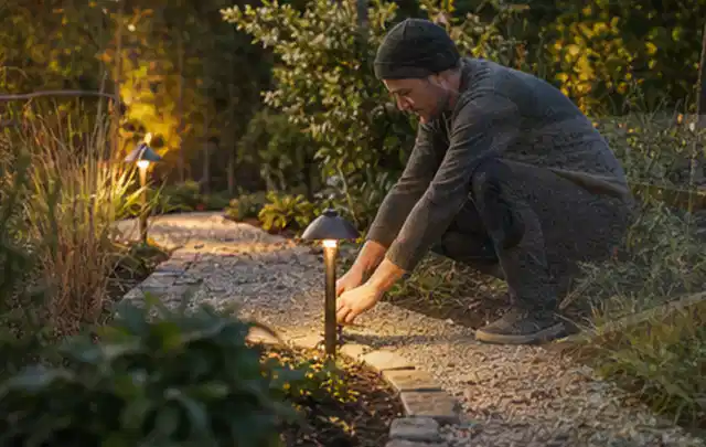 Person richtet eine niedrige warm leuchtende Gartenlampe am Kiesweg aus; Thema Hollywoodschaukeln, Schwerpunkt Kaufberatung; Kontrolle.