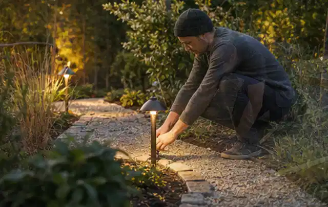 Person richtet eine niedrige warm leuchtende Gartenlampe am Kiesweg aus; Thema Hollywoodschaukeln, Schwerpunkt Nachhaltigkeit; Umsetzung.