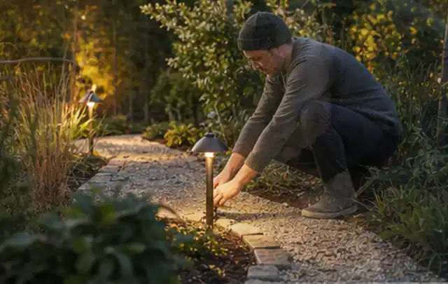 Person richtet eine niedrige warm leuchtende Gartenlampe am Kiesweg aus; Thema Hollywoodschaukeln, Schwerpunkt Testkriterien; Ausgangslage.