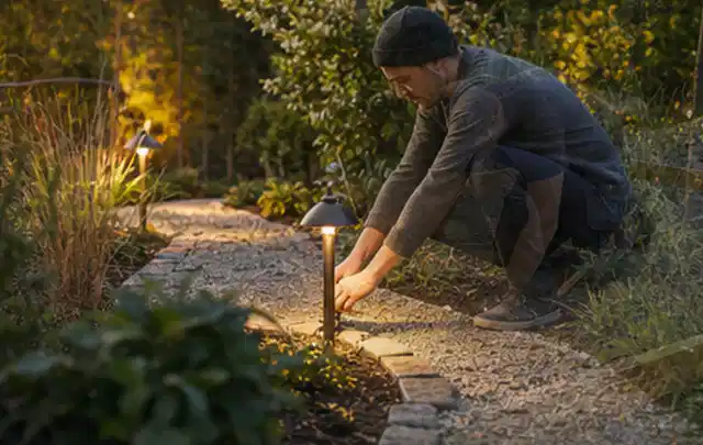 Person richtet eine niedrige warm leuchtende Gartenlampe am Kiesweg aus; Thema Hollywoodschaukeln, Schwerpunkt Winterlager; Ausgangslage.