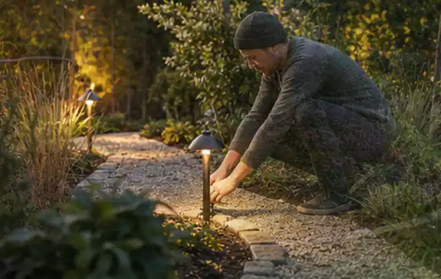Person richtet eine niedrige warm leuchtende Gartenlampe am Kiesweg aus; Thema Holzkohlegrills, Schwerpunkt Anwendung; Praxisalltag.