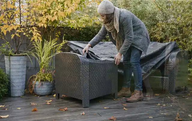 Person deckt Gartenmöbel vor der kalten Jahreszeit sorgfältig ab; Thema Holzkohlegrills, Schwerpunkt Checkliste; Praxisalltag.