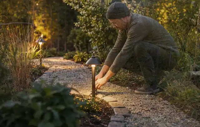 Person richtet eine niedrige warm leuchtende Gartenlampe am Kiesweg aus; Thema Holzkohlegrills, Schwerpunkt Dimensionierung; Ausgangslage.