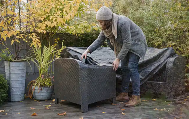Person deckt Gartenmöbel vor der kalten Jahreszeit sorgfältig ab; Thema Holzkohlegrills, Schwerpunkt Dimensionierung; Praxisalltag.