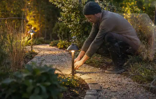 Person richtet eine niedrige warm leuchtende Gartenlampe am Kiesweg aus; Thema Holzkohlegrills, Schwerpunkt Leistung; Kontrolle.
