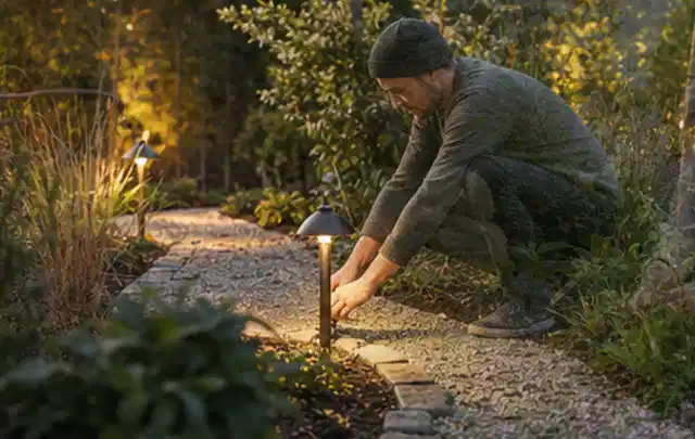 Person richtet eine niedrige warm leuchtende Gartenlampe am Kiesweg aus; Thema Holzkohlegrills, Schwerpunkt Sicherheit; Ausgangslage.