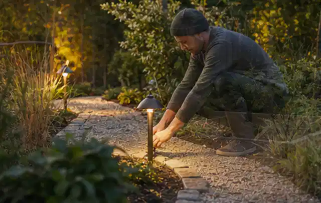 Person richtet eine niedrige warm leuchtende Gartenlampe am Kiesweg aus; Thema Holzzäune, Schwerpunkt Aufbau; Ausgangslage.