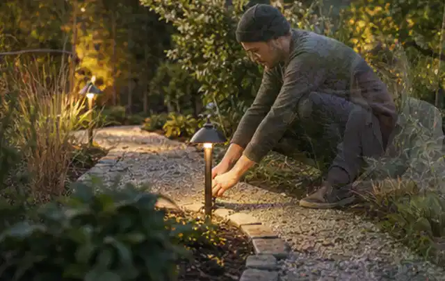 Person richtet eine niedrige warm leuchtende Gartenlampe am Kiesweg aus; Thema Holzzäune, Schwerpunkt Baurecht; Ergebnis.