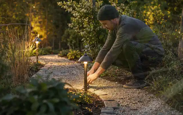 Person richtet eine niedrige warm leuchtende Gartenlampe am Kiesweg aus; Thema Husqvarna Automower, Schwerpunkt Kaufberatung; Ausgangslage.