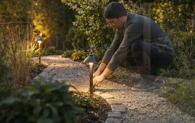 Person richtet eine niedrige warm leuchtende Gartenlampe am Kiesweg aus; Thema Husqvarna Automower, Schwerpunkt Mähroboter; Ausgangslage.