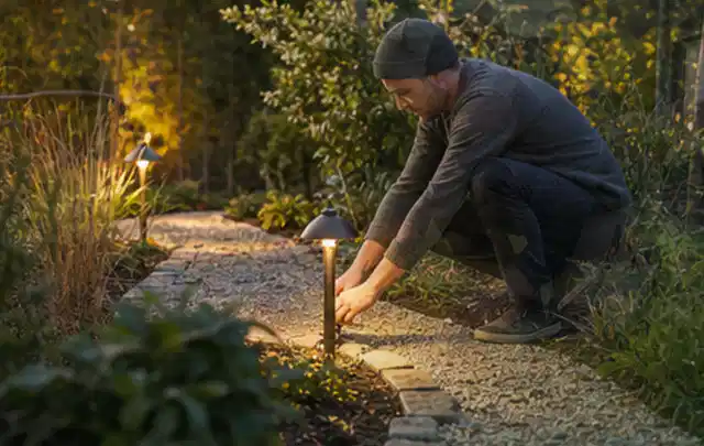 Person richtet eine niedrige warm leuchtende Gartenlampe am Kiesweg aus; Thema Husqvarna Automower, Schwerpunkt Nachhaltigkeit; Umsetzung.