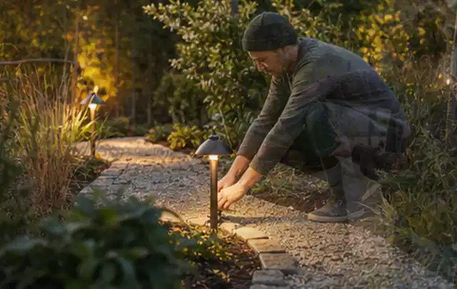 Person richtet eine niedrige warm leuchtende Gartenlampe am Kiesweg aus; Thema Husqvarna Automower, Schwerpunkt Reinigung; Umsetzung.