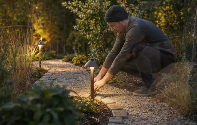 Person richtet eine niedrige warm leuchtende Gartenlampe am Kiesweg aus; Thema Husqvarna Automower, Schwerpunkt Testkriterien; Ergebnis.