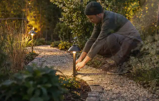 Person richtet eine niedrige warm leuchtende Gartenlampe am Kiesweg aus; Thema Husqvarna Automower, Schwerpunkt Wartung; Kontrolle.
