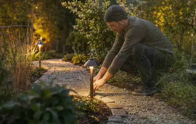 Person richtet eine niedrige warm leuchtende Gartenlampe am Kiesweg aus; Thema Impressum, Schwerpunkt Anbieterkennzeichnung; Umsetzung.