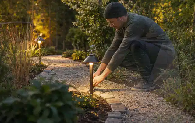 Person richtet eine niedrige warm leuchtende Gartenlampe am Kiesweg aus; Thema Insektenfreundlicher Garten, Schwerpunkt Bepflanzung; Kontrolle.
