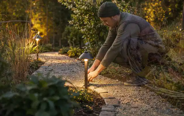 Person richtet eine niedrige warm leuchtende Gartenlampe am Kiesweg aus; Thema Insektenfreundlicher Garten, Schwerpunkt Checkliste; Kontrolle.