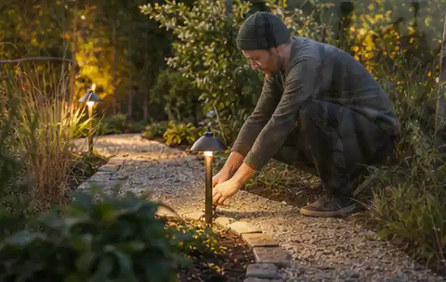 Person richtet eine niedrige warm leuchtende Gartenlampe am Kiesweg aus; Thema Insektenfreundlicher Garten, Schwerpunkt Materialwahl; Praxisalltag.