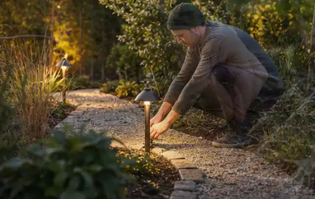 Person richtet eine niedrige warm leuchtende Gartenlampe am Kiesweg aus; Thema Japanischer Garten, Schwerpunkt Kosten; Praxisalltag.